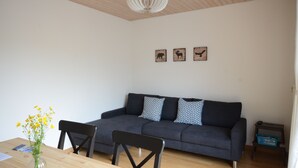Ein modernes Wohnzimmer mit einem dunkelgrauen Sofa, einem Holzespeisetisch mit Stühlen und einer Vase mit gelben Blumen. Darüber hängen drei gerahmte Bilder an der Wand.