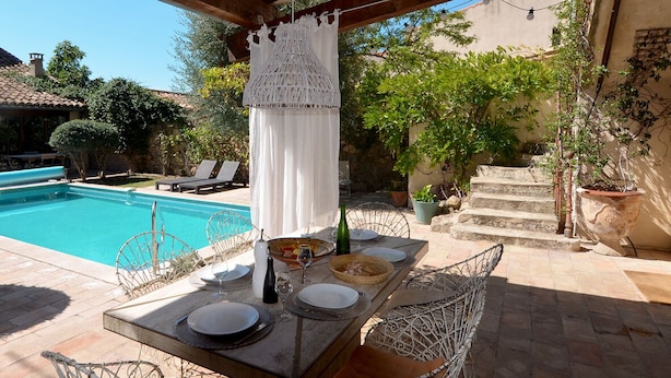 Un comedor junto a la piscina con una mesa para dos, una cortina blanca colgante y una piscina al fondo.