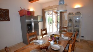 Comedor con mesa de madera dispuesta para cuatro personas, rodeada de sillas. Se ven un refrigerador y un armario con vajilla al fondo.