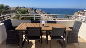 Une terrasse avec une table en bois et des chaises en osier, offrant une vue sur une ville côtière et la mer.