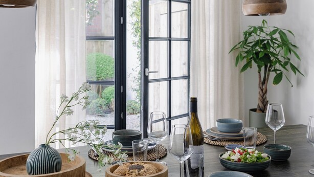 Mesa de comedor dispuesta con platos, tazones, vasos y una botella, con una planta y cortinas al fondo.
