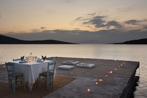 Un comedor con una mesa dispuesta para dos personas, vista al agua y un atardecer de fondo.