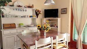 Zona pranzo con tavolo di legno, sedie bianche, un vaso di girasoli, un televisore su un mobile basso e una mensola con diversi oggetti.