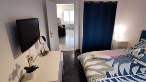 A bedroom with a bed, a TV, a bedside table, a lamp, and a view into the living area. - 1-room apartment for spa guests and vacationers (Balaruc-Les-Bains)