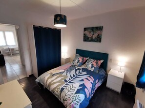 A bedroom with a bed, two bedside tables, a nightstand, a ceiling light, a window with curtains, and a door.