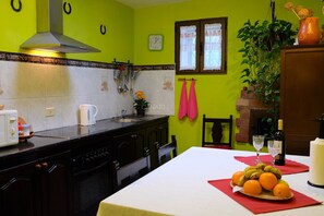 A kitchen with a stove, sink, and cabinets; a dining table with fruit and glasses; a clock on the wall; and a plant.