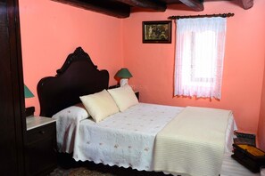 A bedroom with a bed, a nightstand, a lamp, a window with lace curtains, and a picture on the wall.