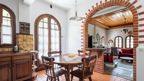 A dining area with a wooden table and chairs, a kitchen with a brick archway, and a red sofa in the background. - Zenios Dionysos - Traditional villa overlooking Pindos (Grevena)