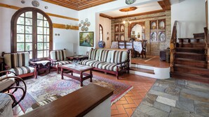 A traditional living room with a wooden ceiling, a large arched window, and a stone fireplace. - Zenios Dionysos - Traditional villa overlooking Pindos (Grevena)