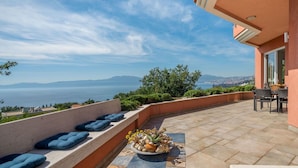 A terrace with a view of the sea, a seating area with cushions, and a table with a flower arrangement.