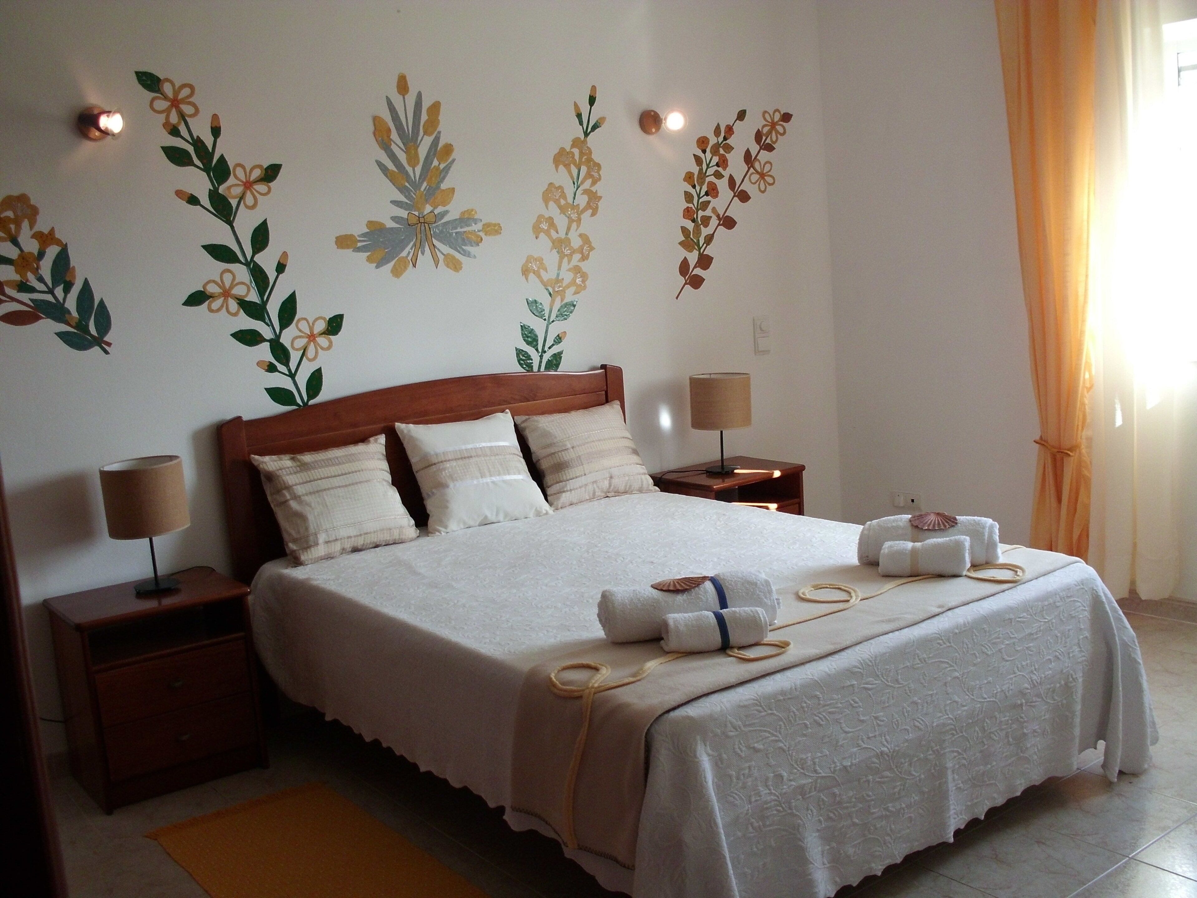 Ein ordentlich bezogenes Bett mit weißer Bettdecke und Kissen, zwei Nachttische mit Lampen und Wandbilder mit floralen Motiven.