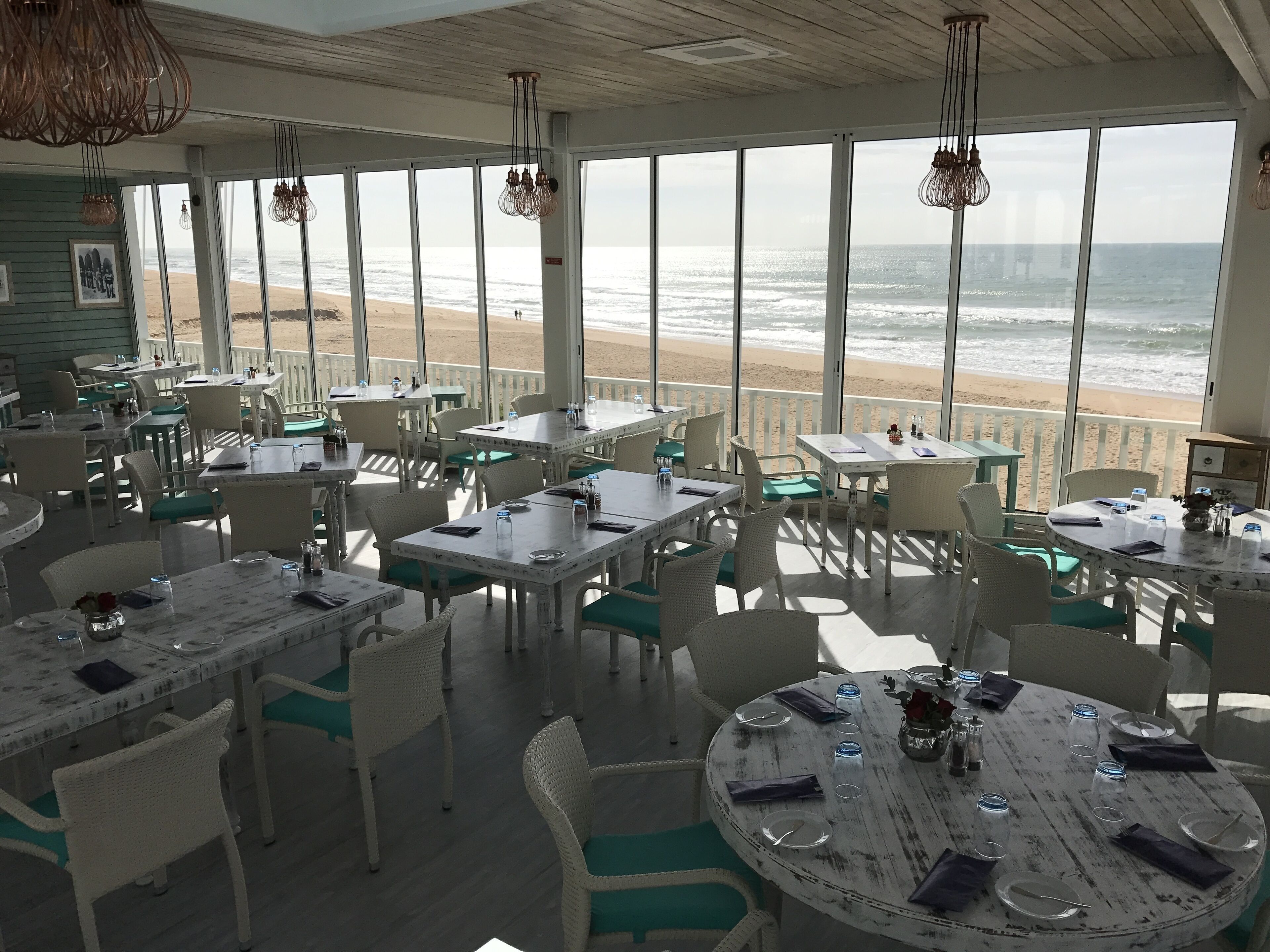 Un restaurant situé en bord de mer, doté d’une terrasse, de grandes fenêtres offrant une vue sur l’océan et d’un décor intérieur rustique.
