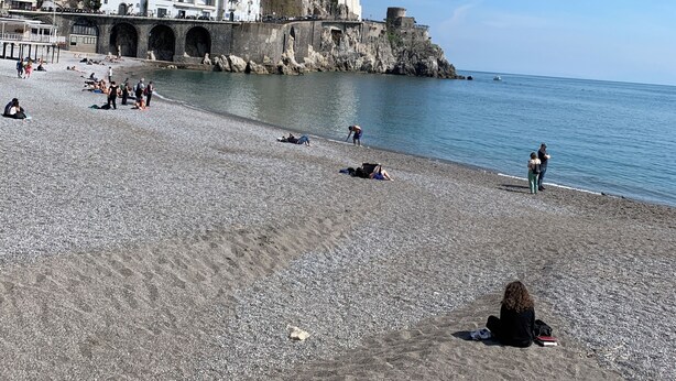 A pebble beach with buildings on a hillside in the background.
