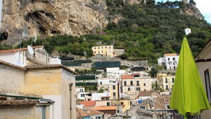 A table with a colorful tablecloth, a green umbrella, and yellow chairs on a terrace overlooking a town with buildings on a hillside.