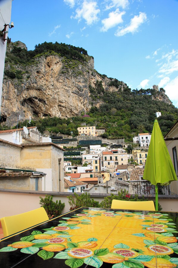 A table with a colorful tablecloth, a green umbrella, and yellow chairs on a terrace overlooking a town with buildings on a hillside.