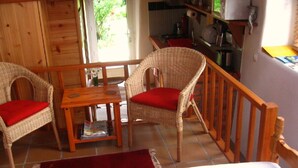 Um quarto aconchegante com cama, mesa de madeira, cadeiras de vime com almofadas vermelhas e uma cozinha visível através de uma porta aberta.