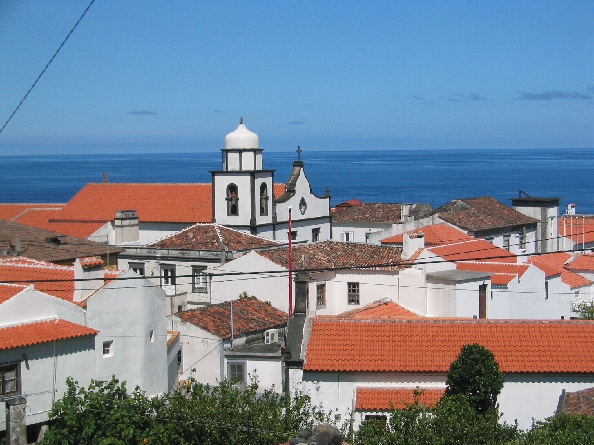 Un pueblo costero con edificios blancos y tejados rojos, una iglesia destacada con un campanario, y el mar al fondo.