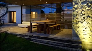 Un patio moderno al aire libre con mesa y bancos de madera, pared de piedra y puerta de cristal que da a un interior blanco.
