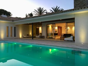Ett modernt hus med en swimmingpool, en täckt uteplats och en glasdörr som leder in till ett inomhusutrymme med en matplats.