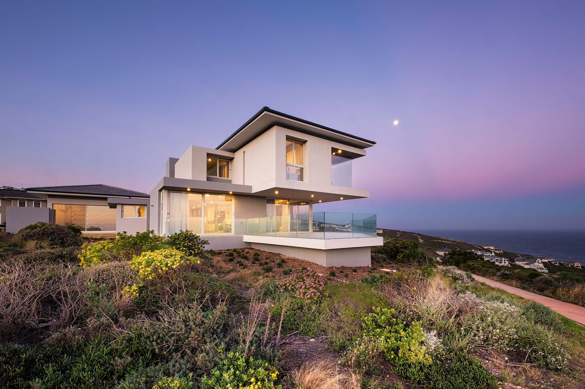 A modern two-story house with a balcony, situated on a hillside with a view of the ocean.