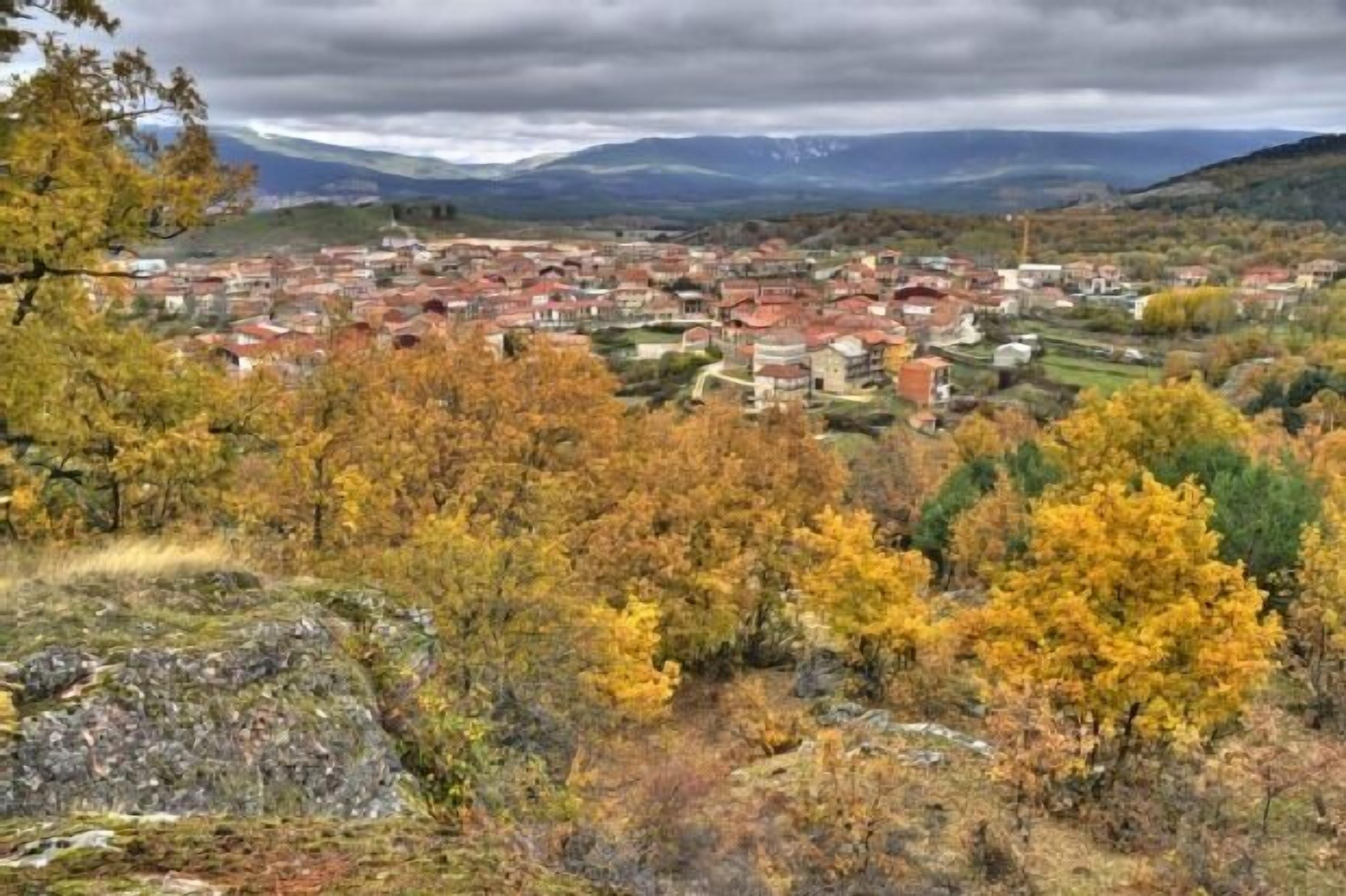 Une ville aux bâtiments aux toits rouges, entourée d’un feuillage automnal et dominée par des montagnes en arrière-plan.