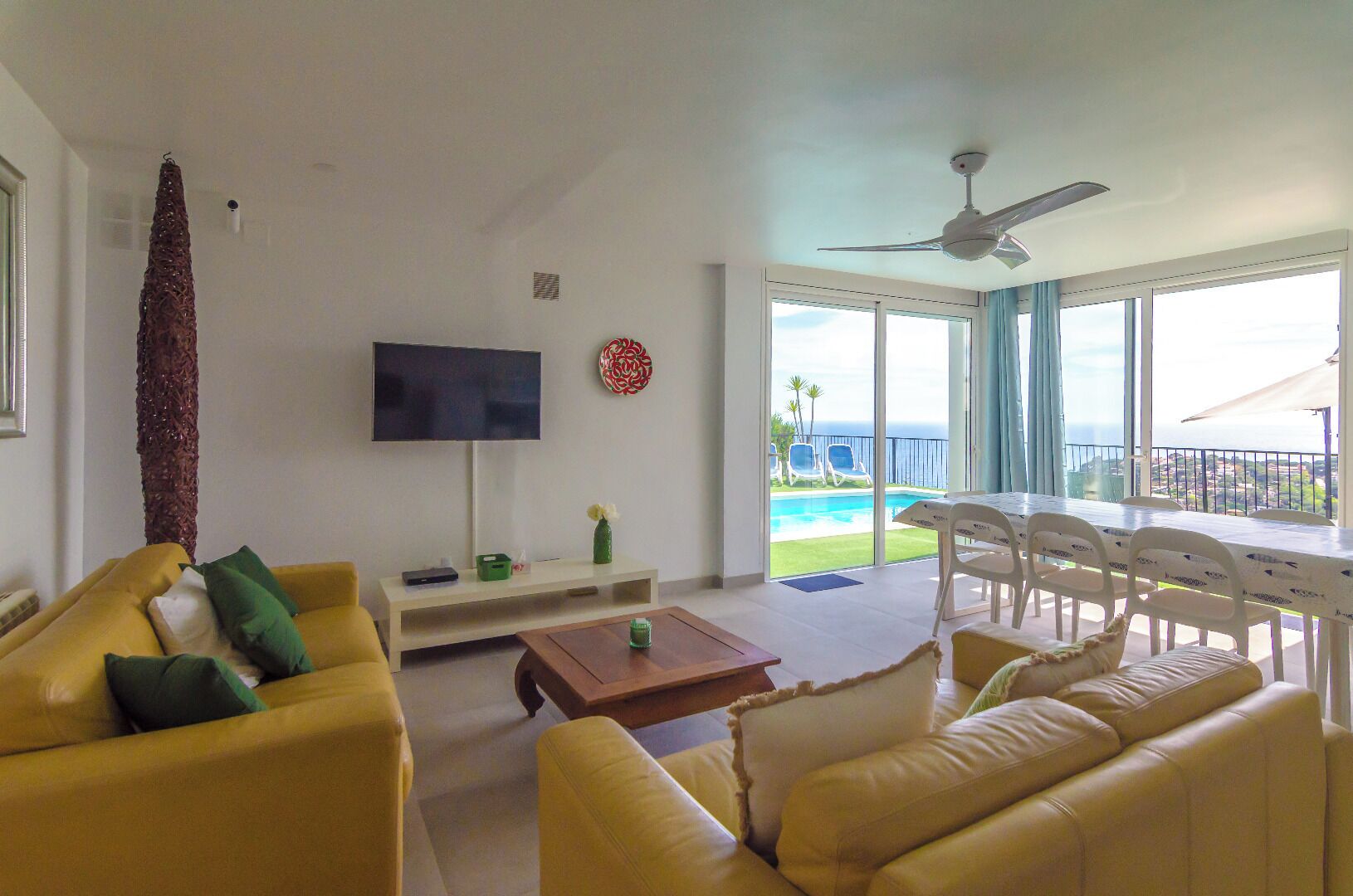 Ein Wohnzimmer mit gelben Sofas, einem Fernseher, einem Deckenventilator und einem Balkon mit Blick auf einen Pool.