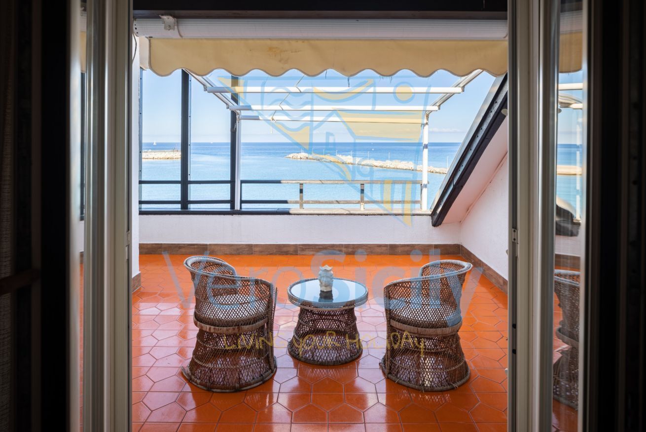 Un balcon avec des fauteuils en osier et une petite table, donnant sur une plage bordée d’une rambarde et offrant une vue sur la mer.