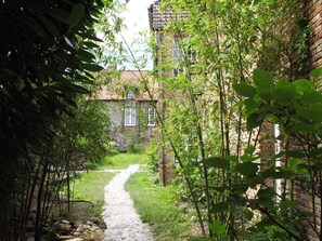 Um caminho de pedra estreito que leva a um edifício de tijolos com várias janelas, rodeado por vegetação exuberante e bambu.