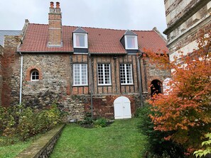 Um edifício de pedra tradicional com telhado em tijolos vermelhos, várias janelas e um pequeno jardim com um caminho que leva à entrada.