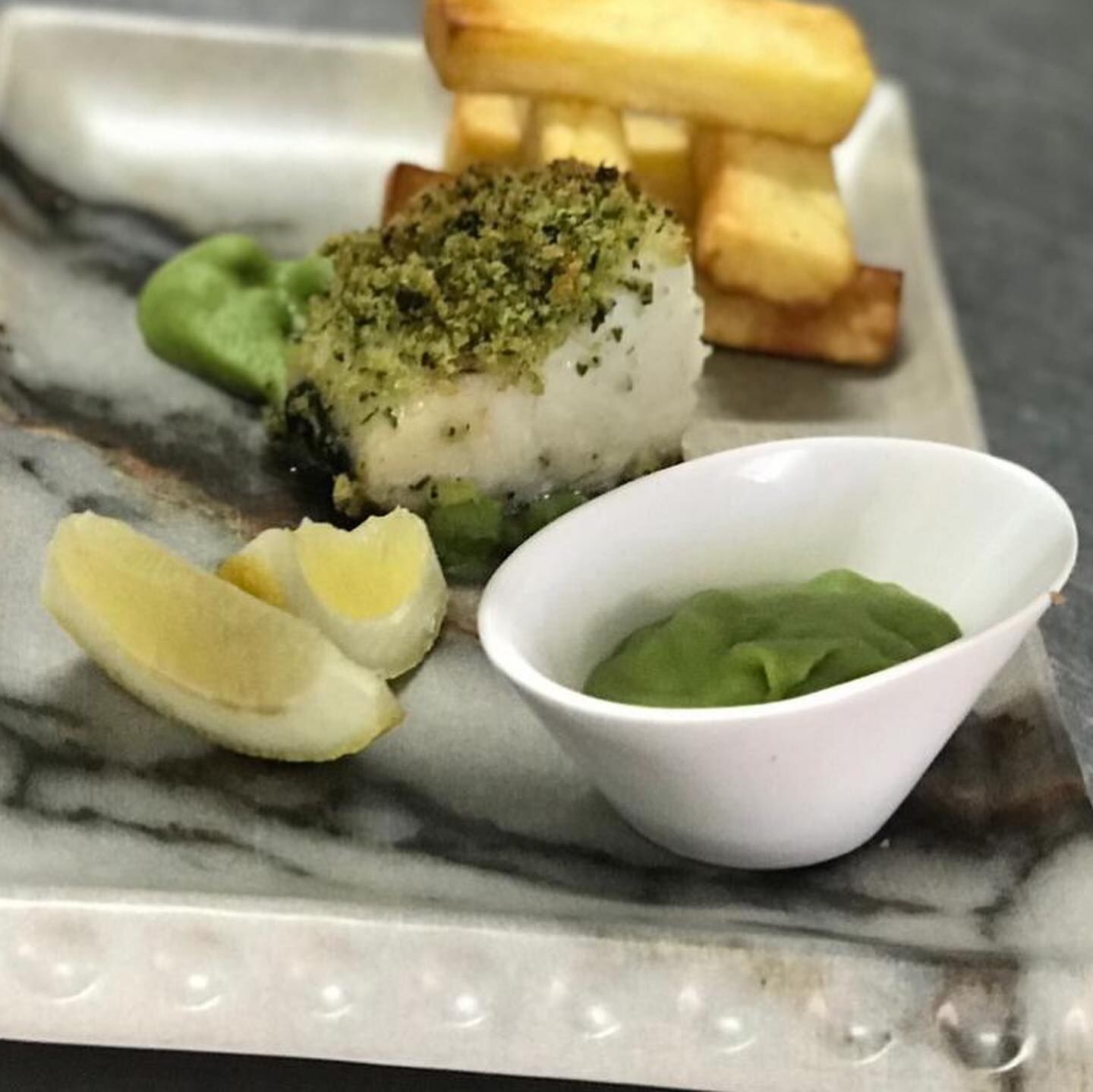 A plate with a piece of fish topped with herbs, two halves of a boiled egg, a small bowl of green sauce, and a side of fried potatoes.