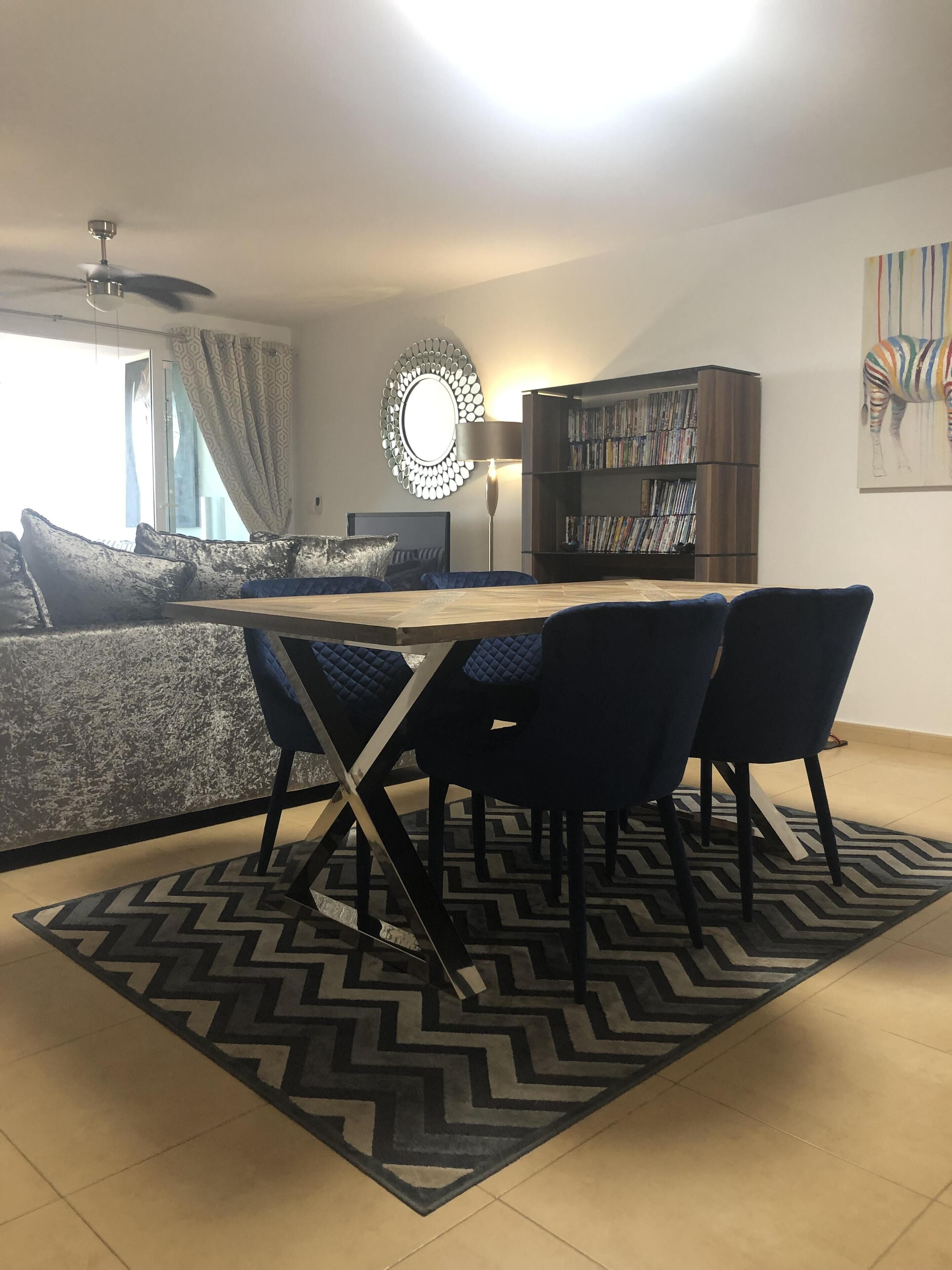 A modern living room with a sofa, a dining table with chairs, a bookshelf, and a ceiling fan.
