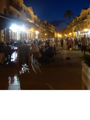 A bustling outdoor dining area with multiple tables and chairs, surrounded by buildings with balconies and palm trees.