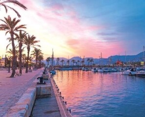 Un port de plaisance avec des bateaux amarrés, une promenade en bord de mer bordée de palmiers et un ciel au coucher du soleil.