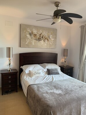 A bedroom with a bed, two bedside tables, a ceiling fan, and a wall-mounted abstract painting.