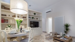 A dining area with a round table, white chairs, and a chandelier. There are bookshelves with books and decorative items. A flat-screen TV is mounted on the wall.