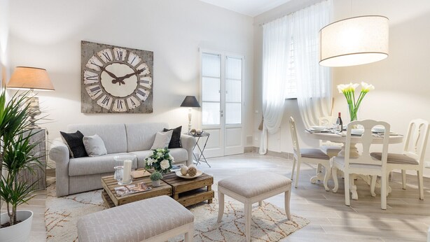 A living room with a sofa, coffee table, and chairs, a dining area with a table and chairs, a large wall clock, and a window with curtains.