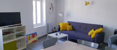 Een woonkamer met een televisie, een bank met gele kussens, een witte salontafel en een boekenkast met boeken en een kleurrijk schilderij.