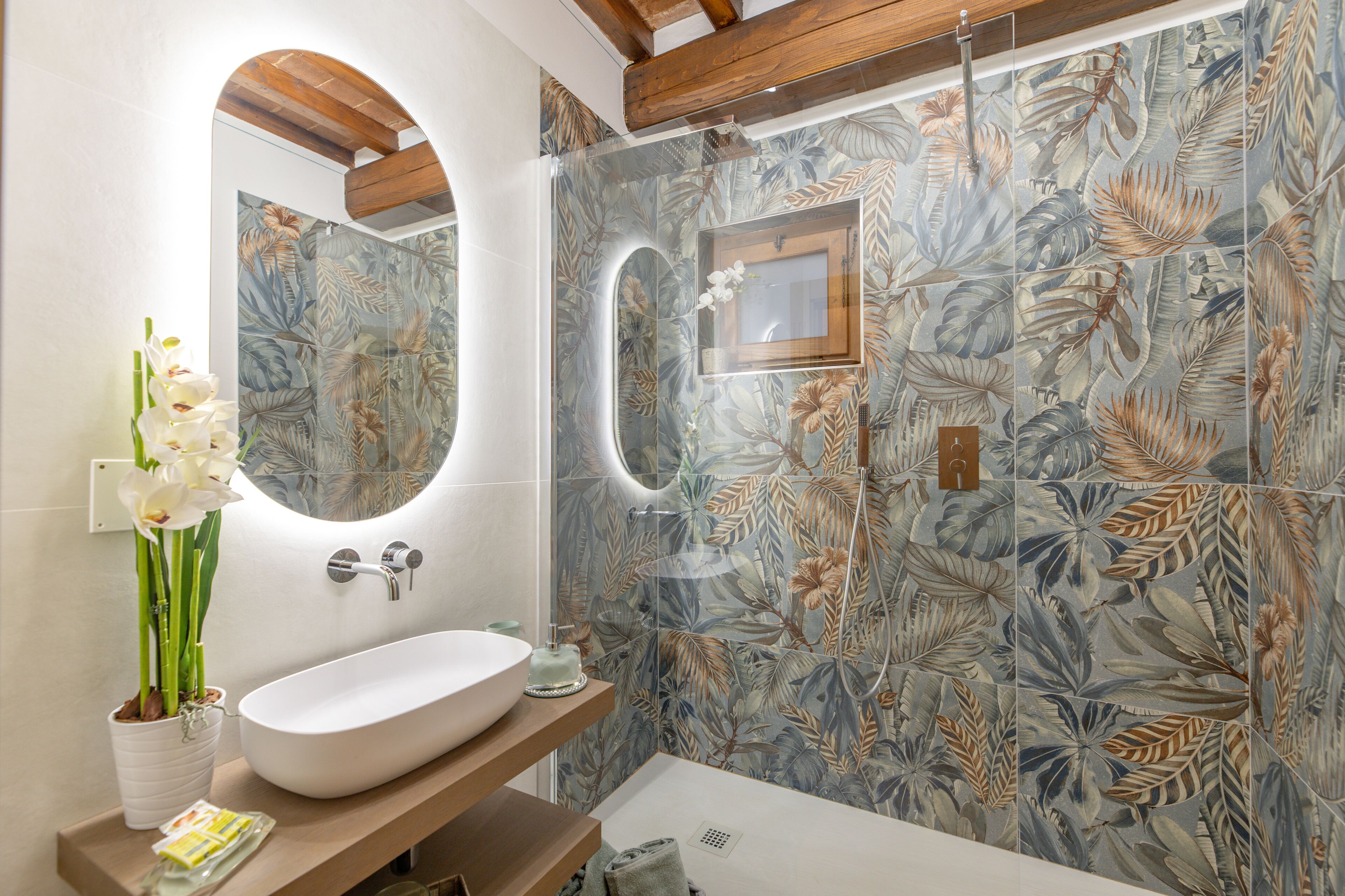 A bathroom with a large oval sink, a mirror, and a shower area with a floral tile design.