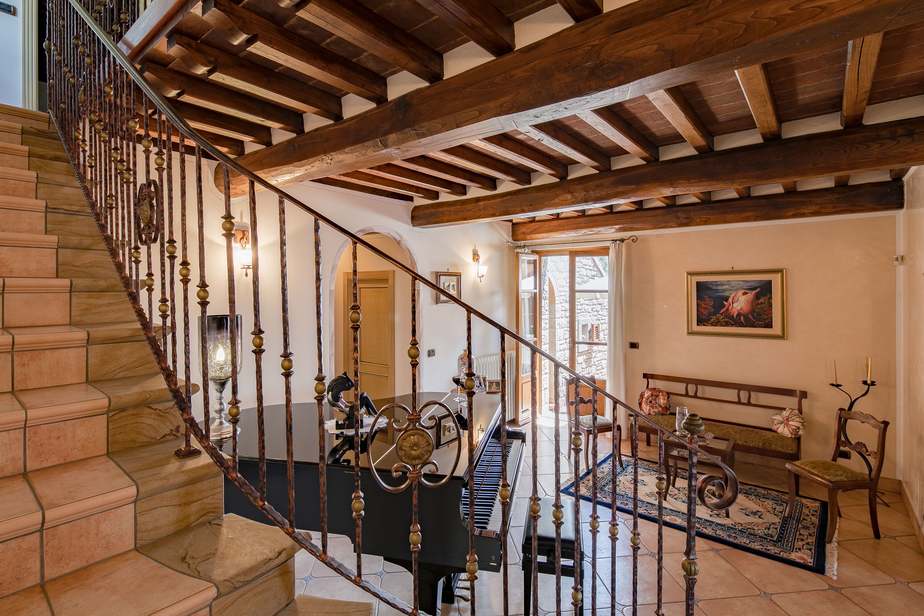 A grand staircase with a wrought iron railing leading to a living area with a piano, a bench, and a painting on the wall.