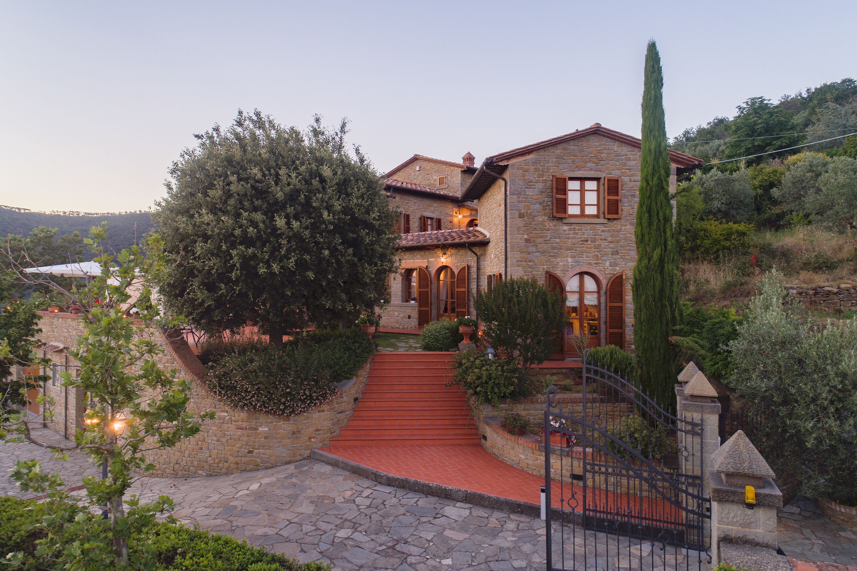 A stone house with a red-tiled roof, a stone staircase leading to the entrance, and a surrounding garden with various plants and a tall tree.