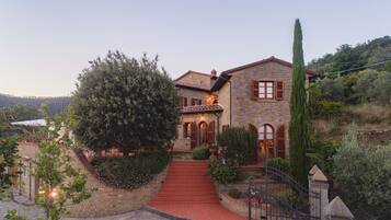 A stone house with a red-tiled roof, a stone staircase leading to the entrance, and a surrounding garden with various plants and a tall tree.