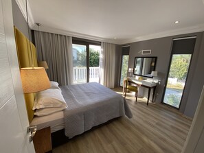 A bedroom with a bed, a desk, a chair, a large window, and a balcony.
