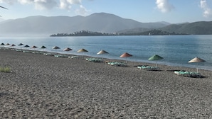 Een kiezelstrand met rijen groene ligbedden en kleurrijke parasols, met uitzicht op een rustige zee en verre bergen.