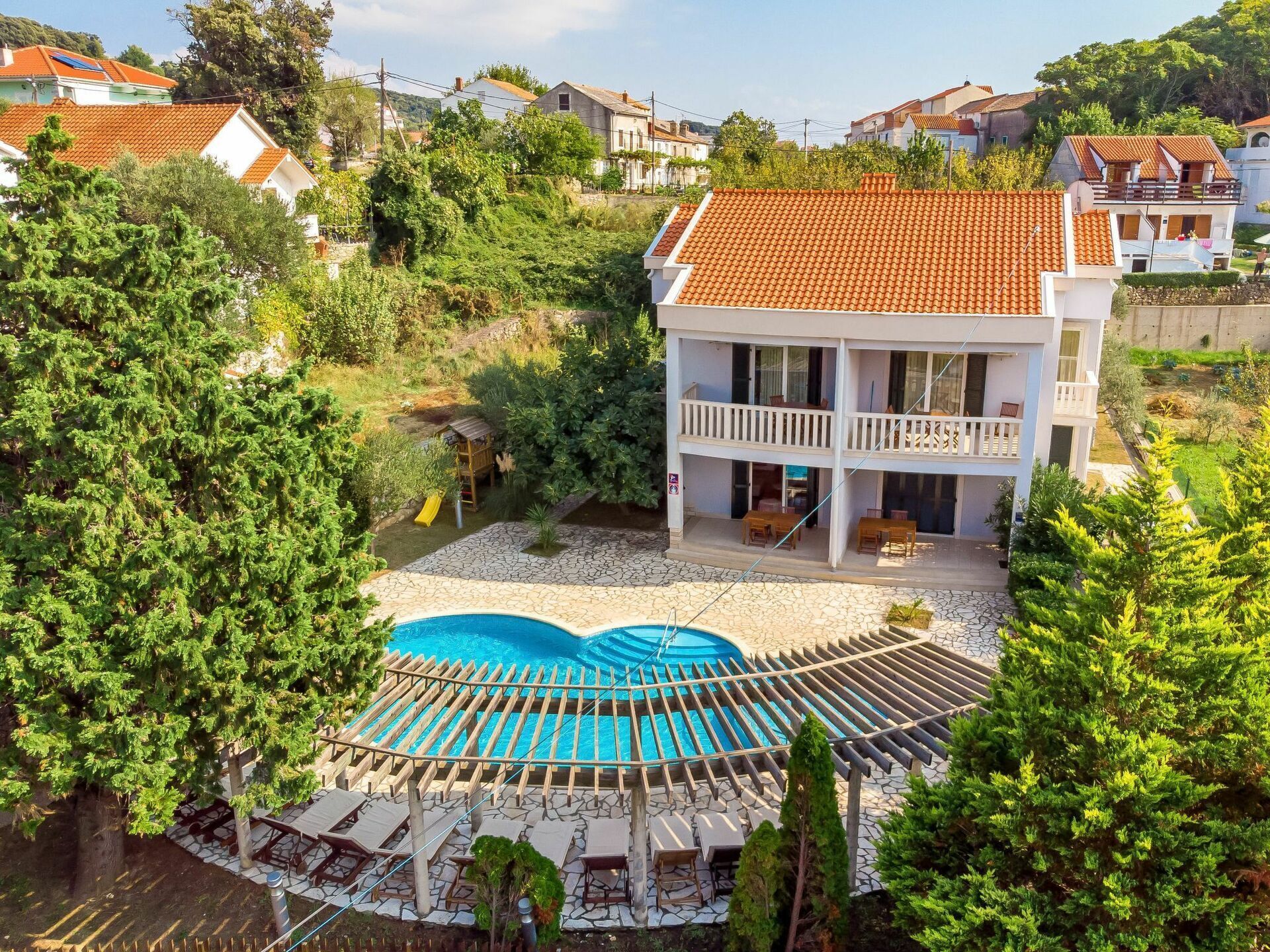 A two-story house with a swimming pool, surrounded by greenery and other houses.