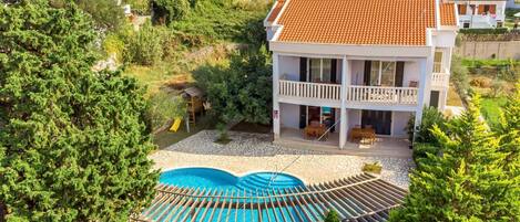 A two-story house with a swimming pool, surrounded by greenery and other houses.
