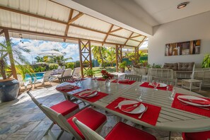 Un espace extérieur aménagé pour les repas, avec un patio couvert, des chaises rembourrées de couleur rouge et une vue sur la piscine.