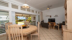 Een ruime woonkamer met een eettafel, stoelen, een bank en een televisie. Er is een balkon met uitzicht op groen.