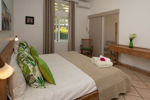 Un lit bien fait, avec un oreiller vert, une couverture beige et une serviette blanche ornée d’une fleur rouge. On y voit également une chaise en bois et une petite table sur laquelle est posé un vase vert.