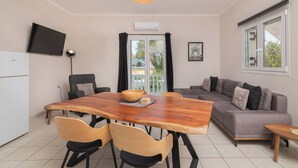 A modern living space with a wooden dining table, two chairs, a grey sofa, a TV mounted on the wall, and a window with curtains.