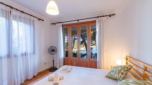 A hotel room with a bed, a fan, a window with curtains, and a view of the outdoors. - Paraiso sobre el Mar (Urbanización de la Torre, Porto Petro, Santanyí)